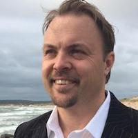 A man with short light brown hair, a goatee, and a moustache, wearing a white shirt and dark jacket, is smiling whilst standing outdoors near the sea on a cloudy day.