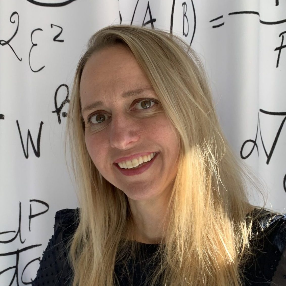A woman with long blonde hair smiles at the camera. She is standing in front of a white background with black mathematical symbols and equations drawn on it.