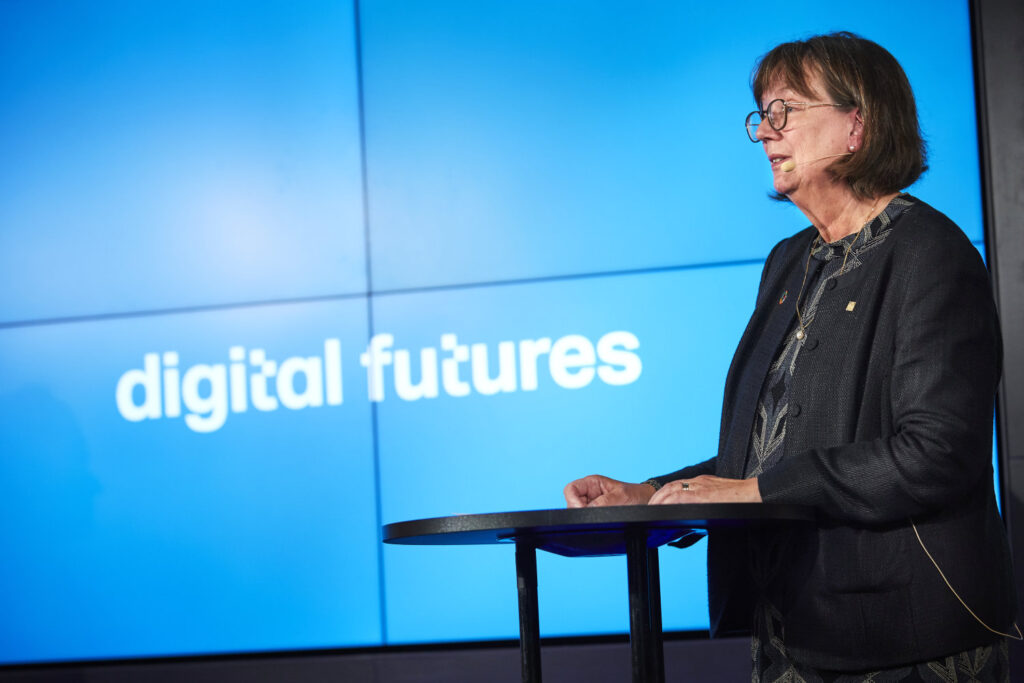 A woman wearing glasses and a dark suit speaks at a lectern. Behind her is a large blue screen displaying the words digital futures.