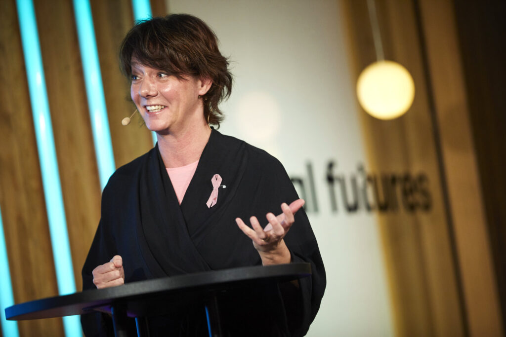 A woman wearing a black outfit and a pink ribbon speaks at a lectern, gesturing with her hand. She smiles, with blurred lights and vertical blue lines in the background.
