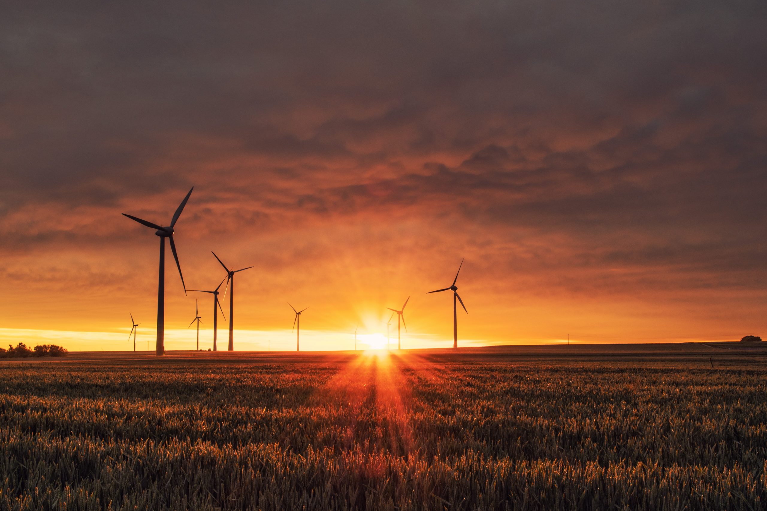 Wind turbines stand in a field under a dramatic, cloudy sky with the sun setting on the horizon, casting an orange glow over the landscape.