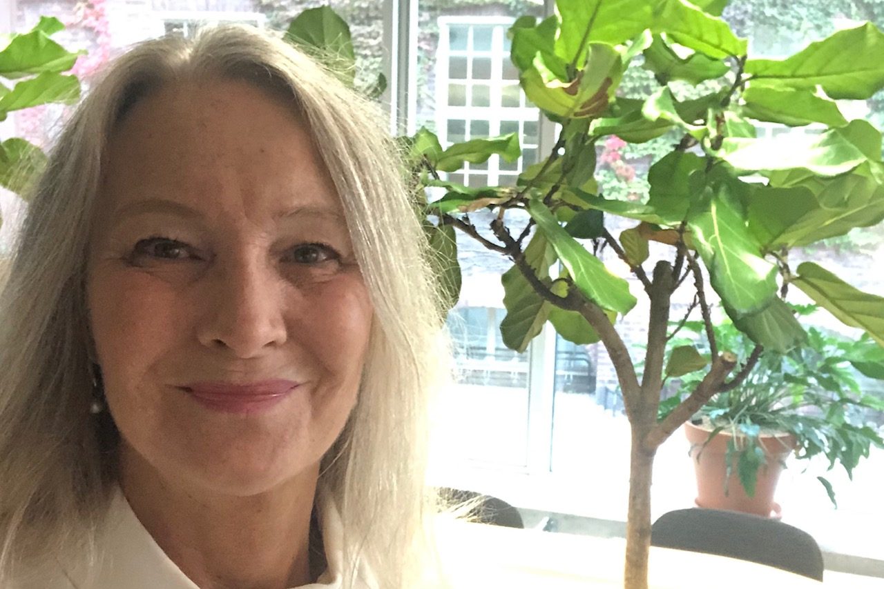 Smiling older woman with long grey hair stands indoors next to a potted fiddle-leaf fig plant, with sunlight coming through large windows and more greenery visible outside.