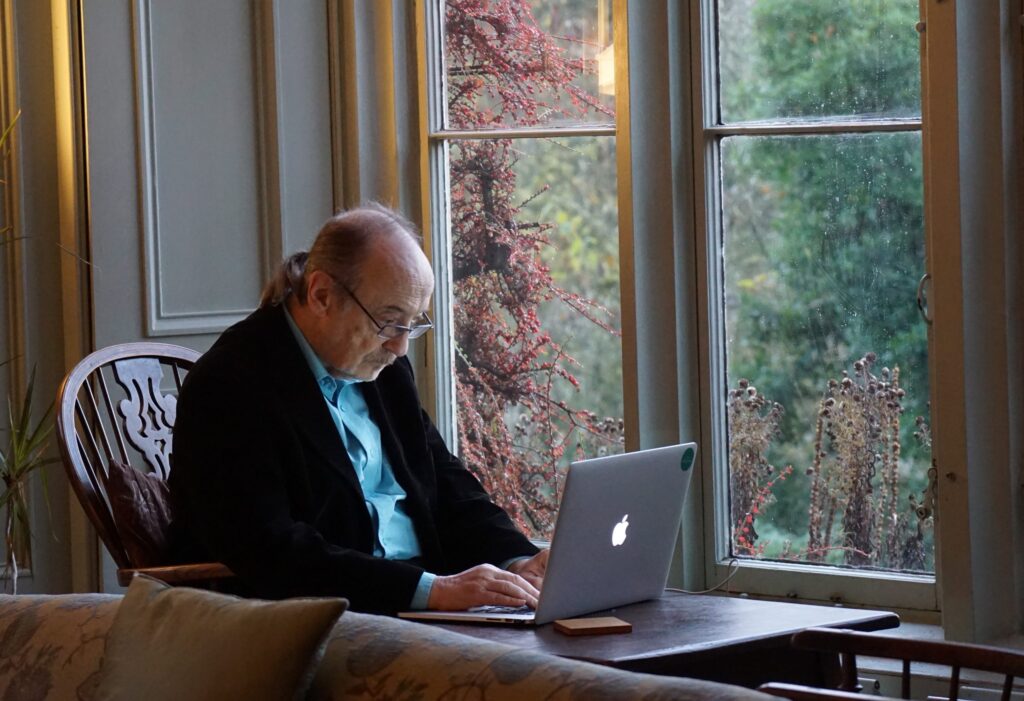 An older man with glasses sits at a wooden table, typing on a laptop near a large window with a garden view. Natural light fills the room, highlighting his focused expression.