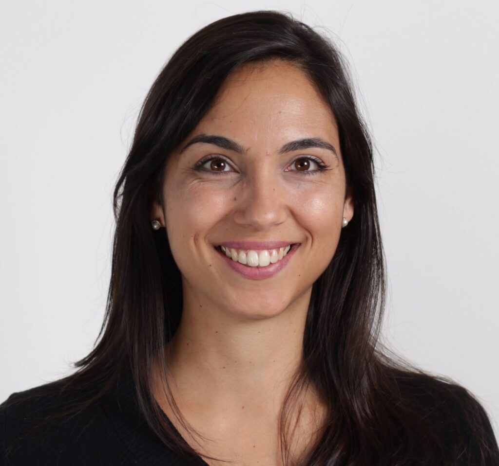 A woman with long dark hair, wearing a black top, smiles at the camera against a plain white background.