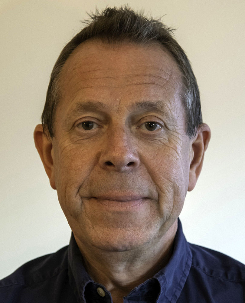 A middle-aged man with short brown hair and a slight smile, wearing a dark blue collared shirt, photographed against a plain light background.