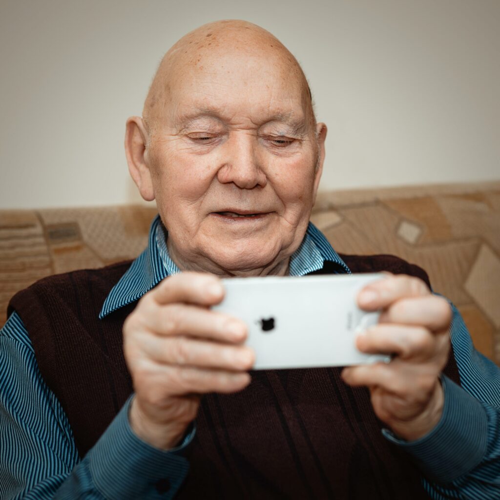 An elderly man with a bald head sits on a sofa, smiling and looking at a mobile phone he is holding in both hands. He wears a blue striped shirt and dark jumper.