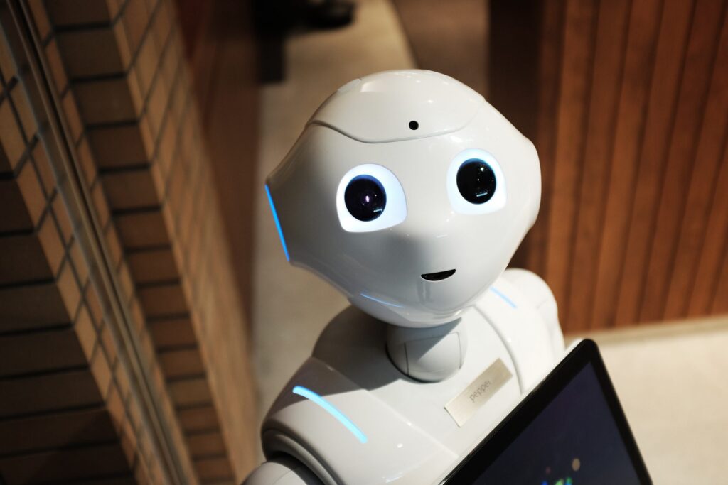 A white humanoid robot with big, glowing eyes and a small smile stands indoors near wood and brick walls, holding a tablet-like device.
