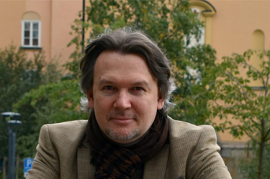 A middle-aged man with wavy grey hair and a short beard wears a brown jacket and scarf, standing outdoors with trees and a peach-coloured building in the background.