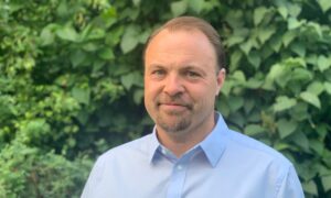 A man with light brown hair and a trimmed beard, wearing a light blue collared shirt, stands outside in front of dense green foliage, smiling slightly at the camera.