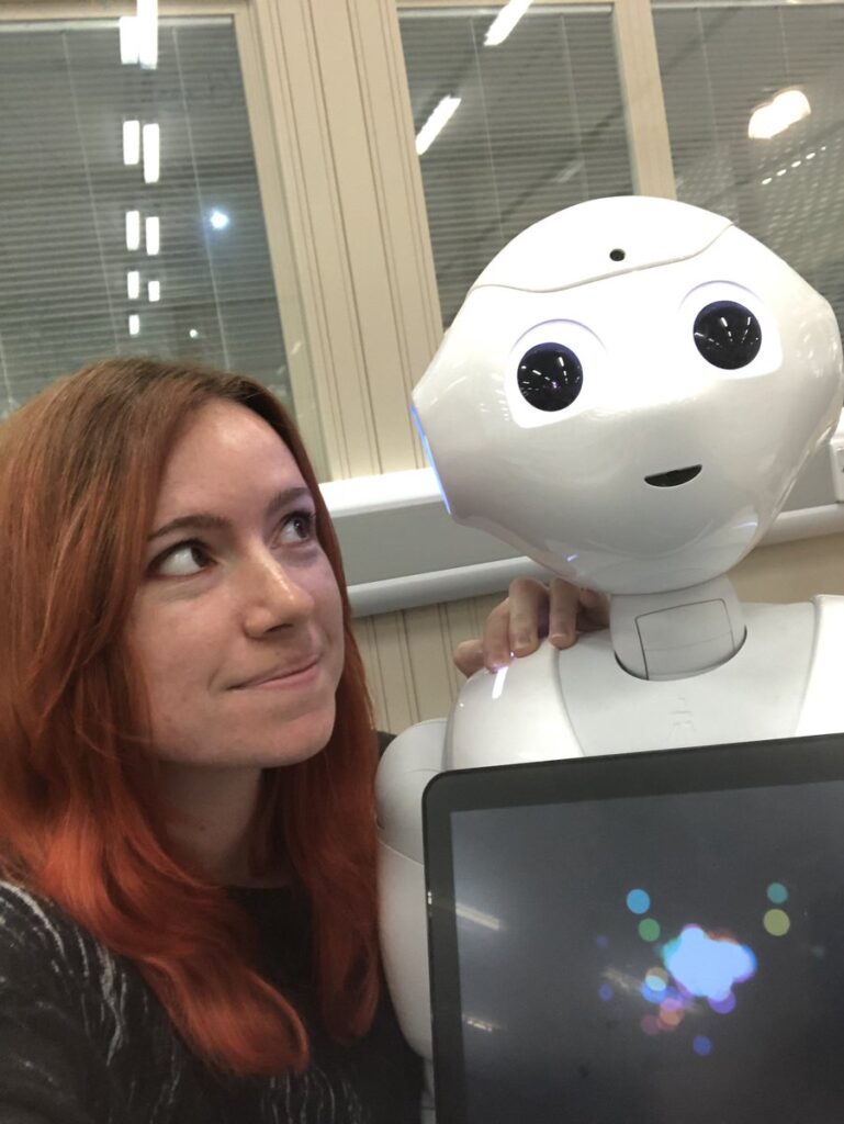 A person with long red hair smiles and looks at a white humanoid robot with large round eyes. The robot has a screen on its chest displaying colourful lights, and they appear to be in an indoor setting with windows behind them.