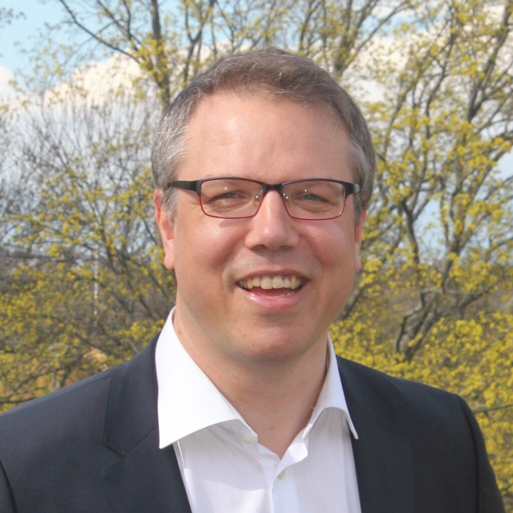 A man with short light brown hair, wearing glasses, a white dress shirt, and a dark suit jacket, smiles outdoors with trees and blue sky in the background.