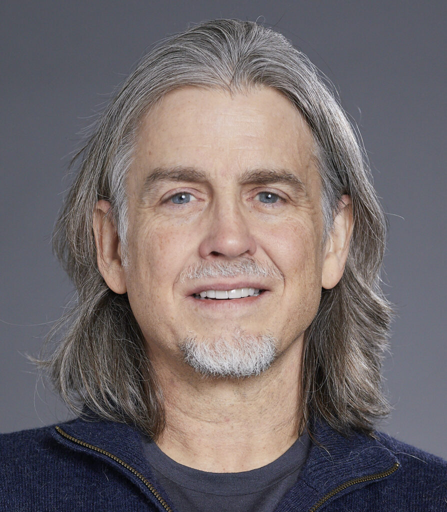 A middle-aged man with shoulder-length grey hair, light facial hair, and blue eyes is smiling gently at the camera. He is wearing a dark zip-up jumper over a dark shirt, with a plain grey background behind him.