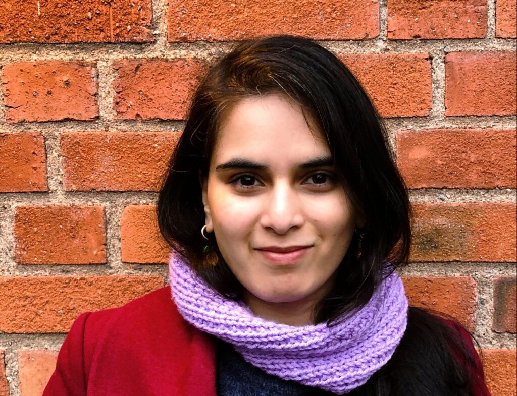 A woman with long dark hair, wearing a maroon coat and a light purple knitted scarf, stands in front of a red brick wall and smiles gently at the camera.