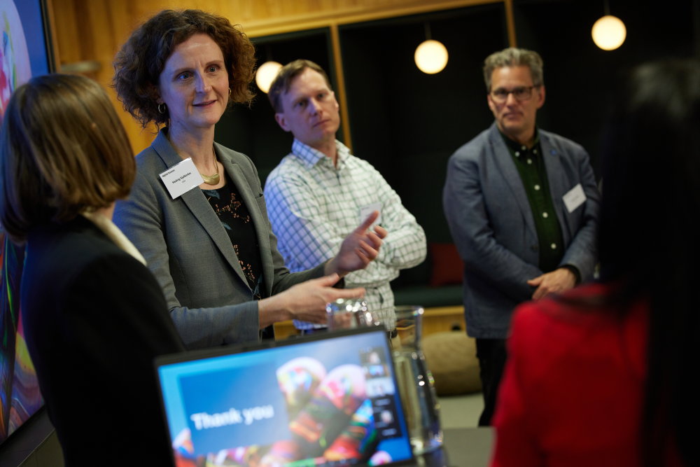 Four professionally dressed adults stand and converse around a table in a modern office setting. A blurred screen in the foreground displays “Thank you.” Three people face the camera, one has their back turned.