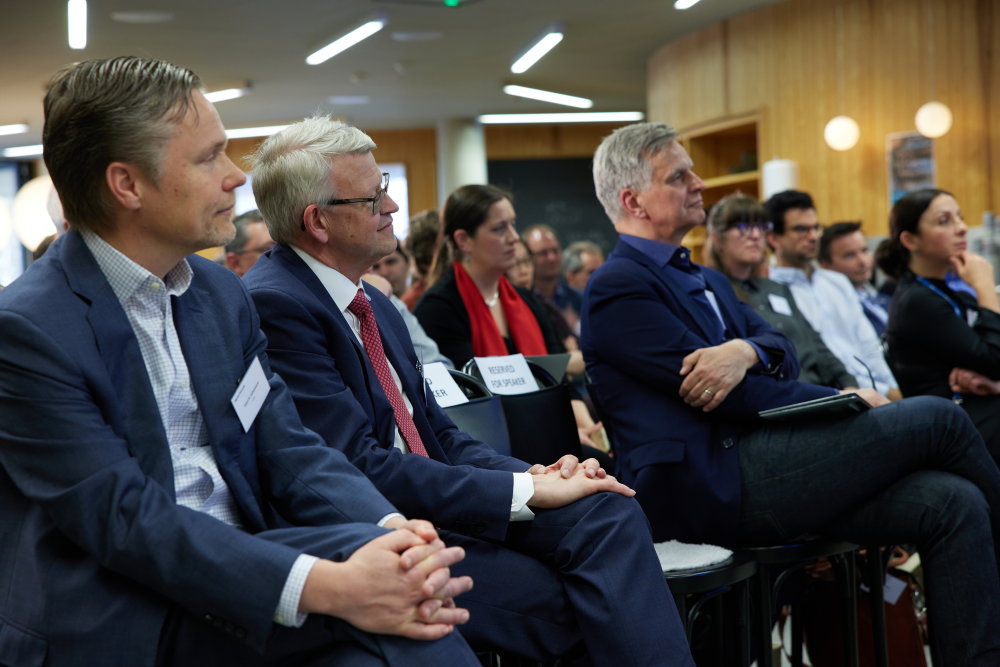 Audience of professionally dressed men and women sit attentively in a modern conference room with wooden walls, listening to a speaker not visible in the image.