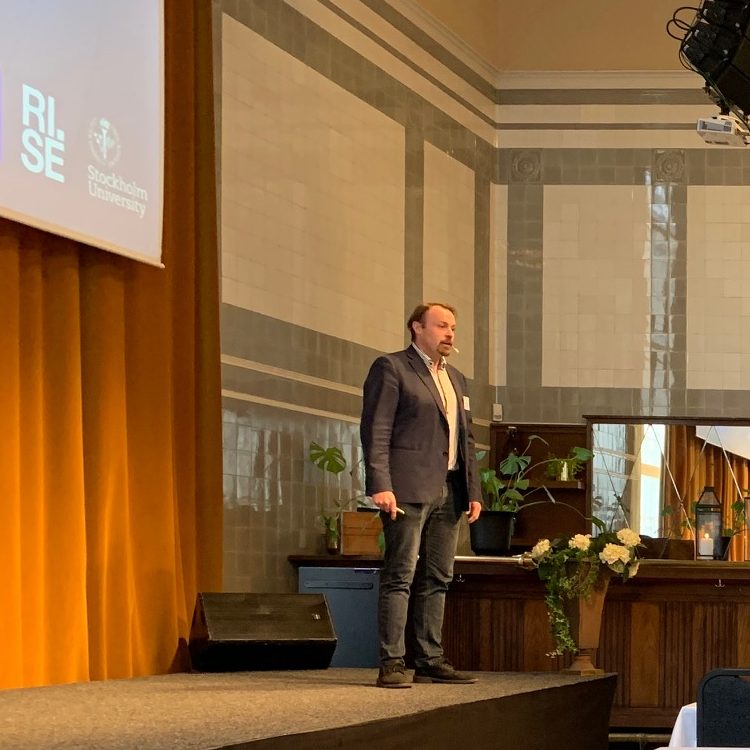 A man in a suit stands on a stage giving a presentation. Behind him are a yellow curtain, plants, and a large screen displaying the RISE and Stockholm University logos. The room has tiled walls and a high ceiling.