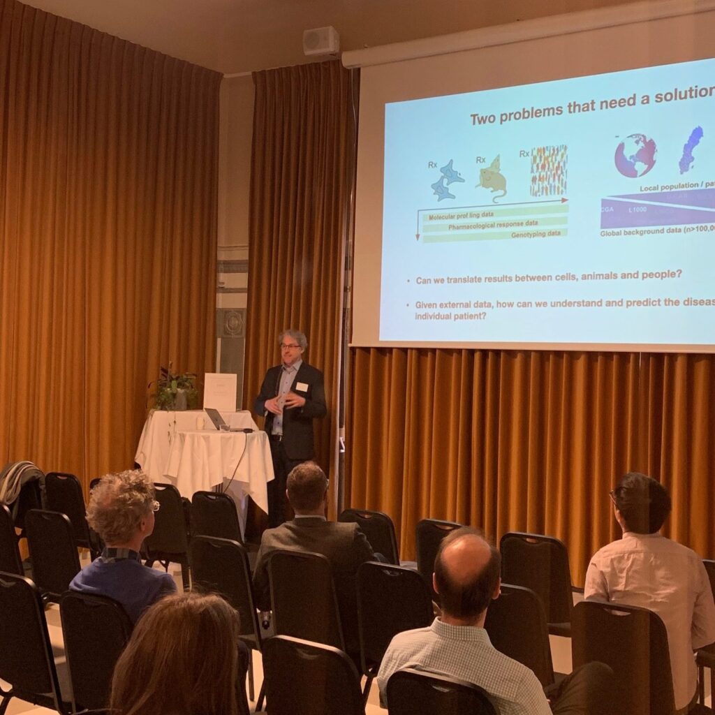 A man gives a presentation in a conference room with brown curtains. He stands at a lectern in front of a screen displaying scientific slides, whilst an audience sits in rows of chairs facing him.