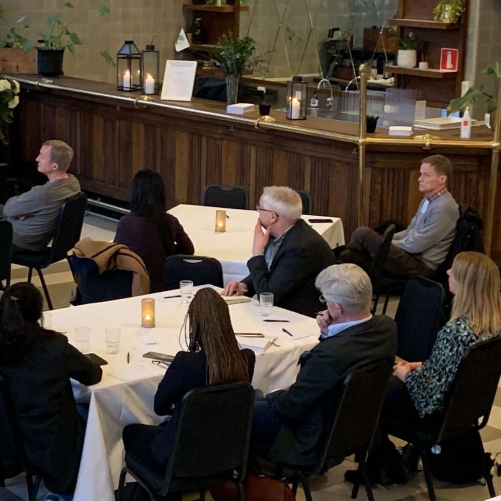 People seated at round tables in a restaurant or café, attentively listening to someone off-camera. Some have notepads and glasses in front of them. The atmosphere is casual and relaxed, with candles and plants decorating the space.