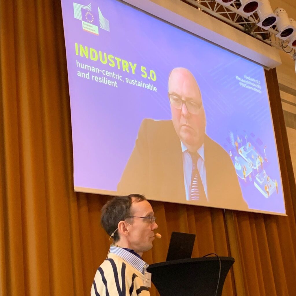A man in glasses speaks at a lectern, while a large screen behind him displays a video call with another man in a suit and the text: INDUSTRY 5.0 human-centric, sustainable and resilient.