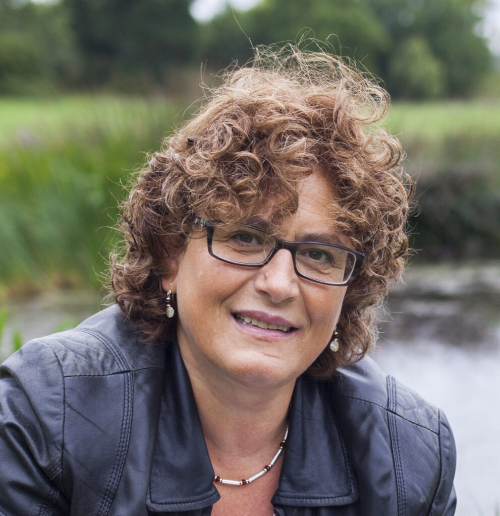 A woman with curly brown hair and glasses, wearing a black leather jacket and silver jewellery, smiles outdoors in front of a pond and greenery.