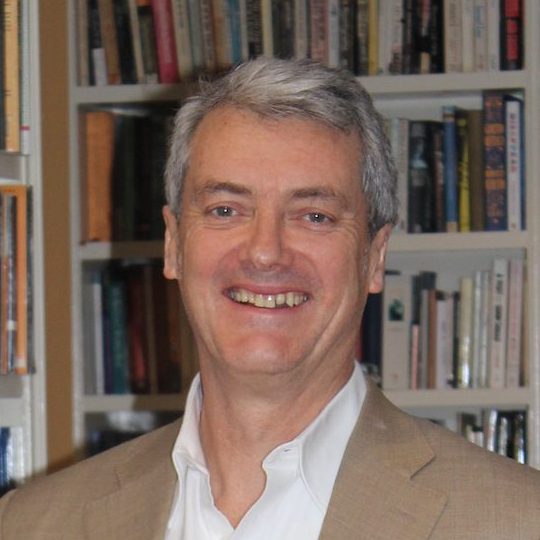 A man with grey hair wearing a beige suit jacket and white shirt smiles in front of a bookcase filled with books.