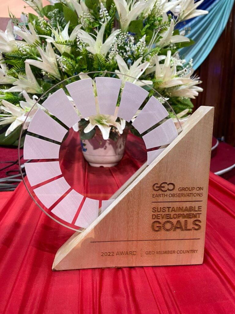 A clear and wooden triangular award trophy with Group on Earth Observations Sustainable Development Goals 2022 Award GEO Member Country engraved, displayed on a red tablecloth with white flowers in the background.