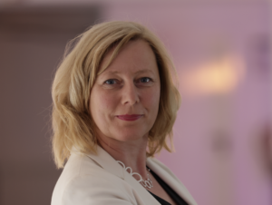 A woman with blonde hair, wearing a white blazer and a necklace, stands in front of a softly blurred background, looking confidently at the camera.