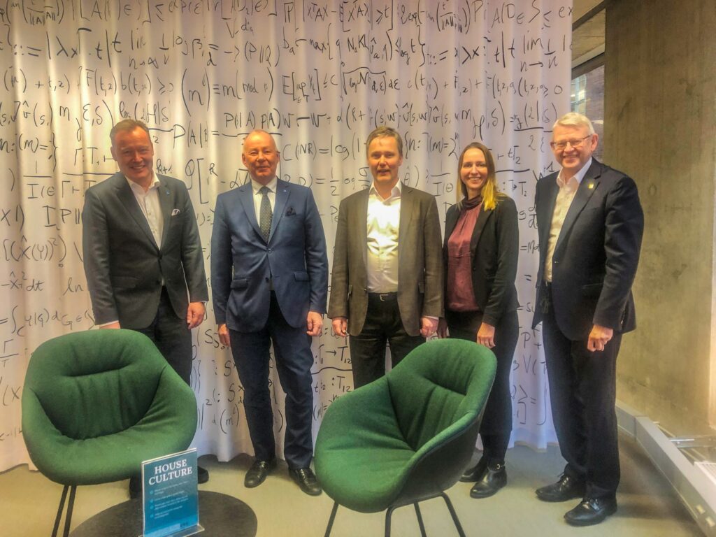 Five people in business attire stand smiling in front of a white curtain covered with mathematical equations. Two green chairs and a small table with a sign reading House of Cultures are in the foreground.