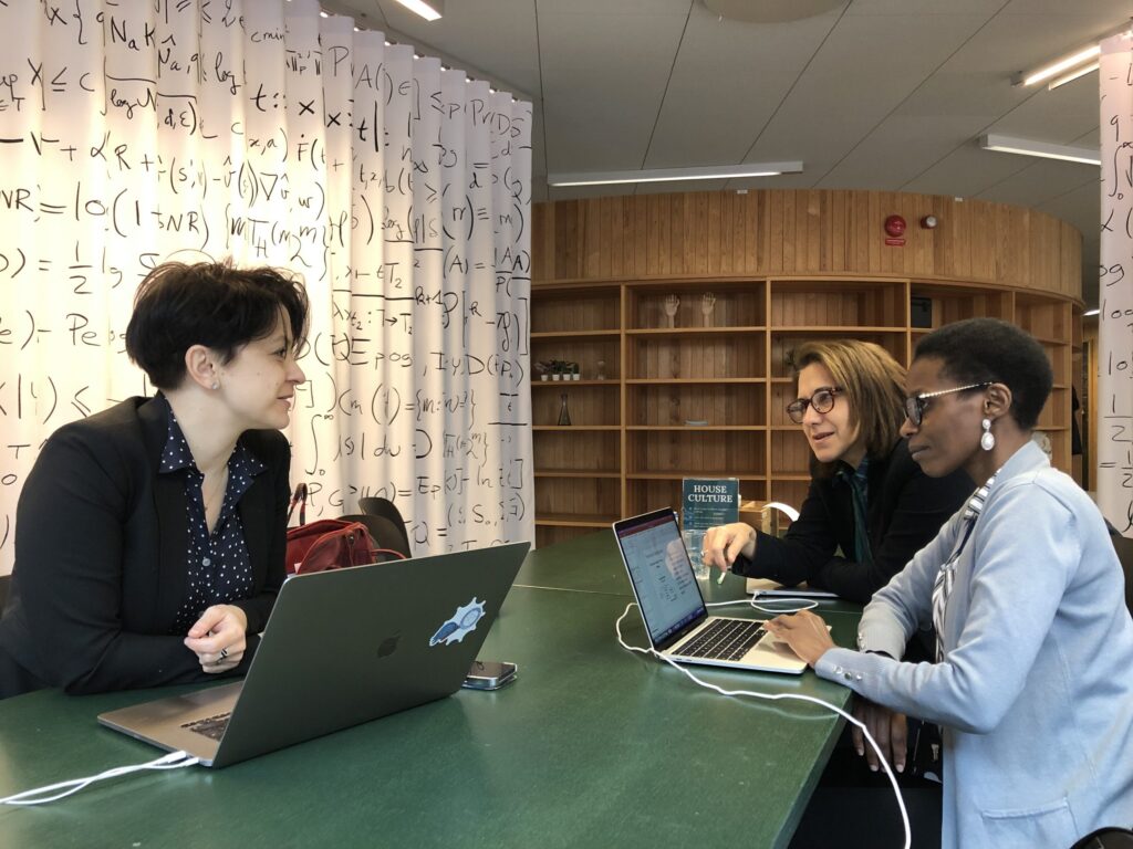 Three women sit at a green table with laptops, engaged in conversation. Behind them are white curtains with mathematical formulae and empty wooden shelves. The setting appears to be a collaborative workspace or study area.