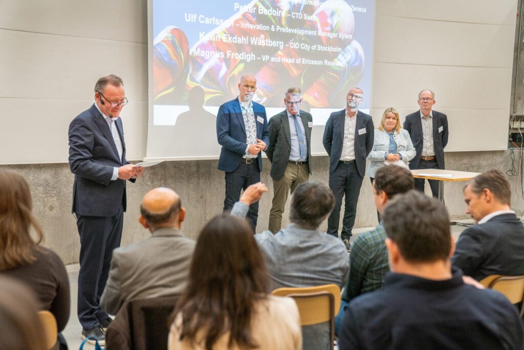 A man stands and speaks to an audience while five people stand together onstage in front of a projected presentation slide at a professional event. Some members of the audience are raising their hands.
