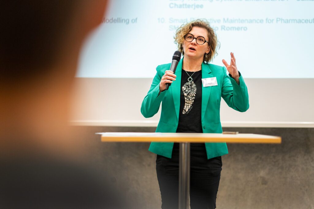 A woman wearing a green jacket speaks into a microphone whilst standing at a round table, gesturing with her hand. A projected presentation with text is visible in the background.