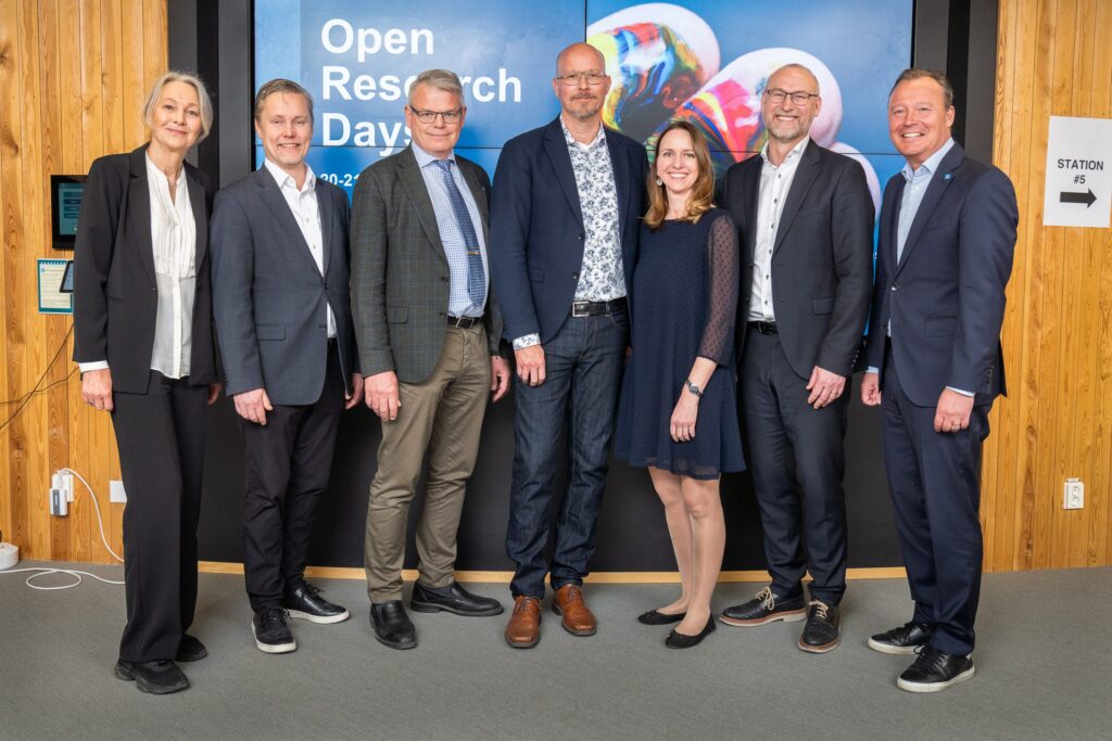 Seven people dressed in business attire stand in a row, smiling, in front of a screen displaying Open Research Day and colourful abstract graphics. The background features wooden walls and a sign reading Station No. 5.