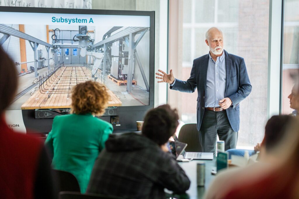 A man in a suit gestures while presenting to a group in a conference room. A large screen behind him displays an image of machinery labelled Subsystem A. Several people are seated and listening attentively.