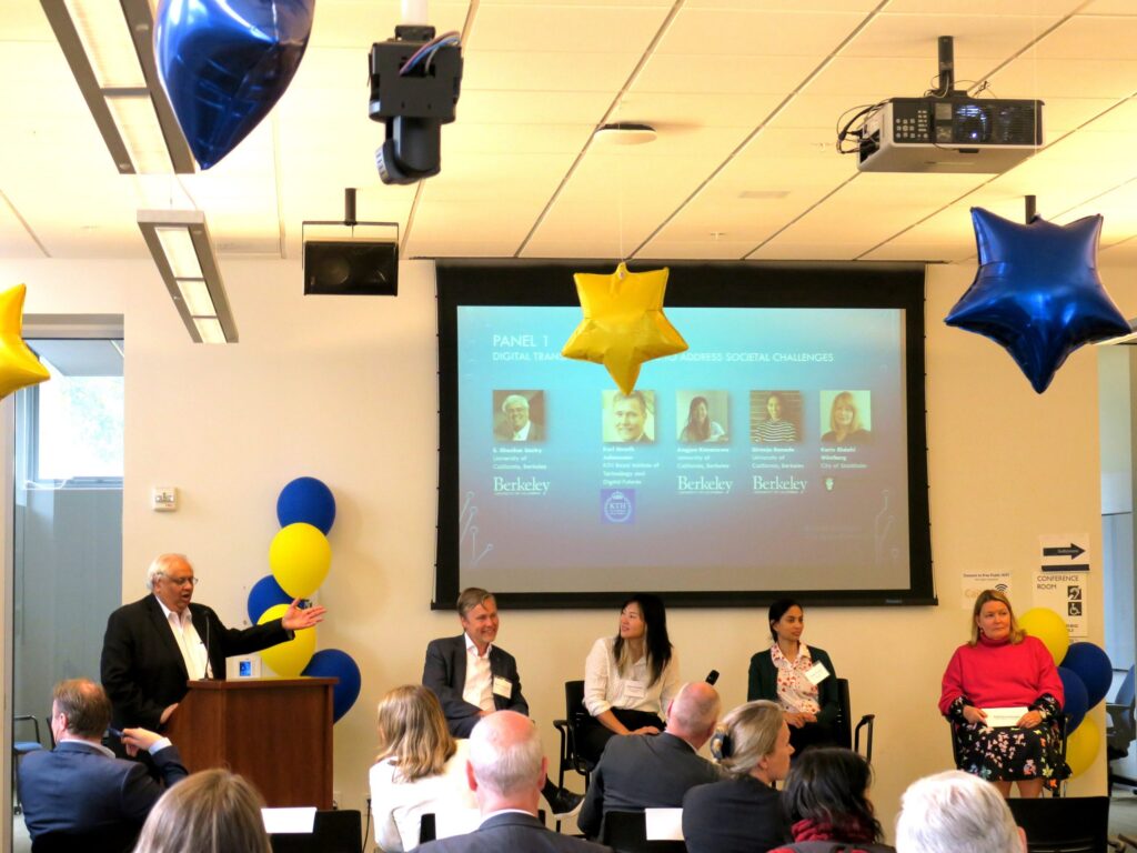 A speaker addresses an audience at a panel event, with four seated panellists beside him and a large display screen behind them. Blue and yellow balloons and star-shaped decorations hang in the room.