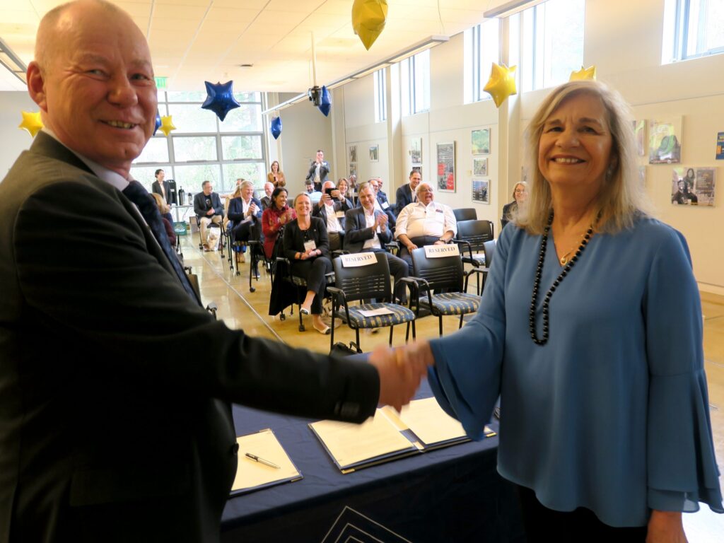 A man and woman smiling and shaking hands at the front of a room, with people seated and applauding in the background. The room is decorated with yellow and blue star-shaped balloons.