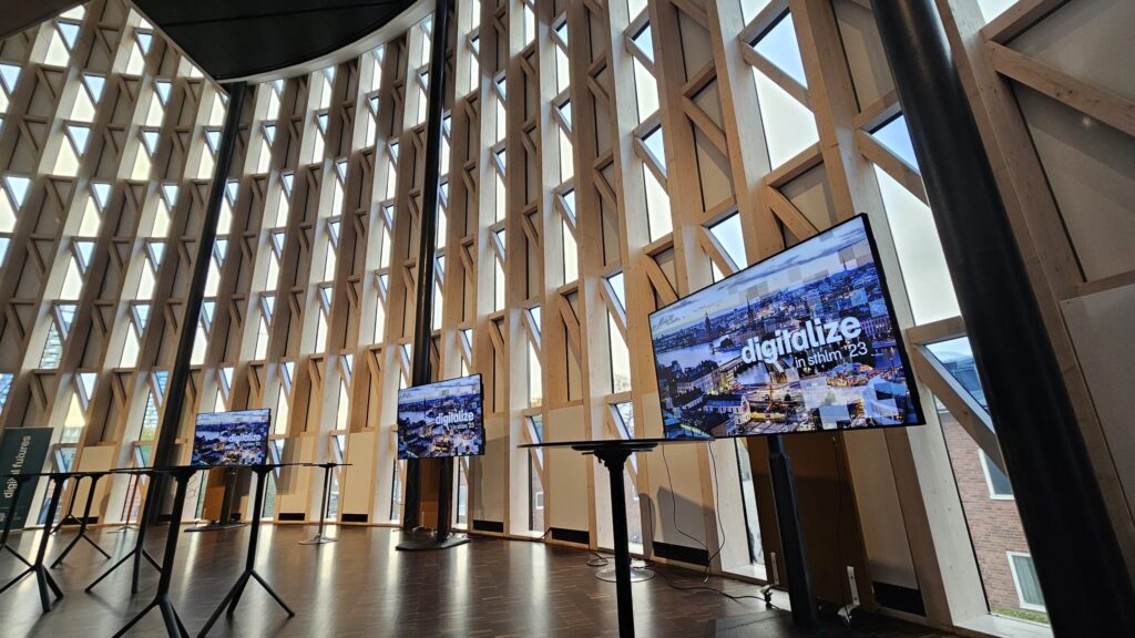 A modern room with tall, wooden grid windows letting in ample sunlight. Three large screens display “digitalise Trondheim 23” with an urban cityscape image beneath the text. Several tall tables line the space.