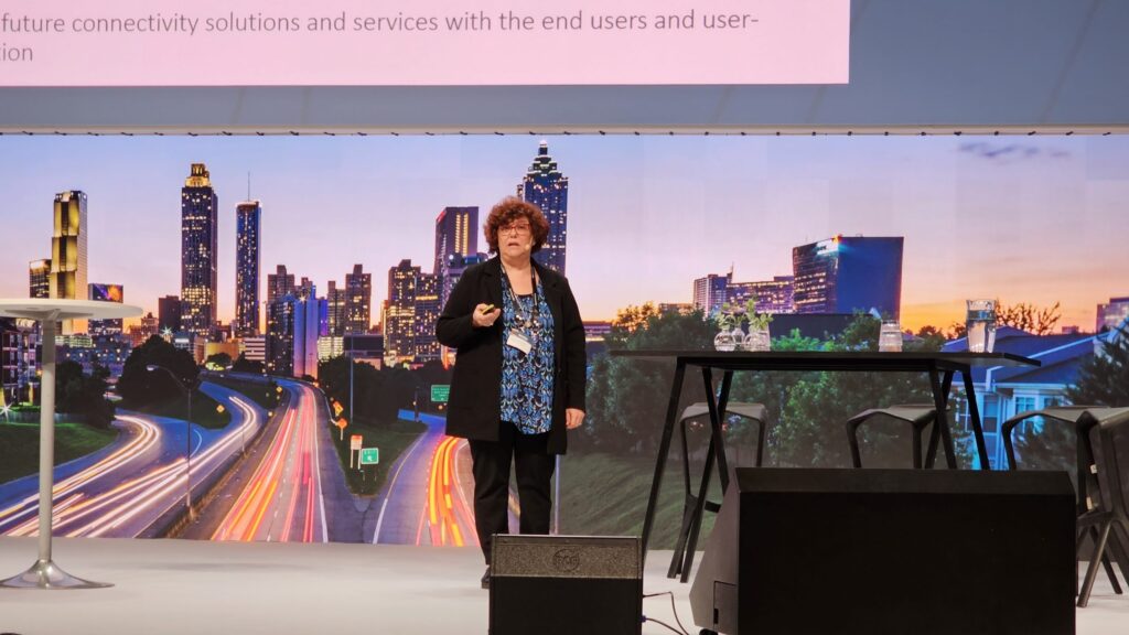 A woman stands on a stage giving a presentation, with a cityscape backdrop and a projected slide above her. There are tables and chairs on the stage.
