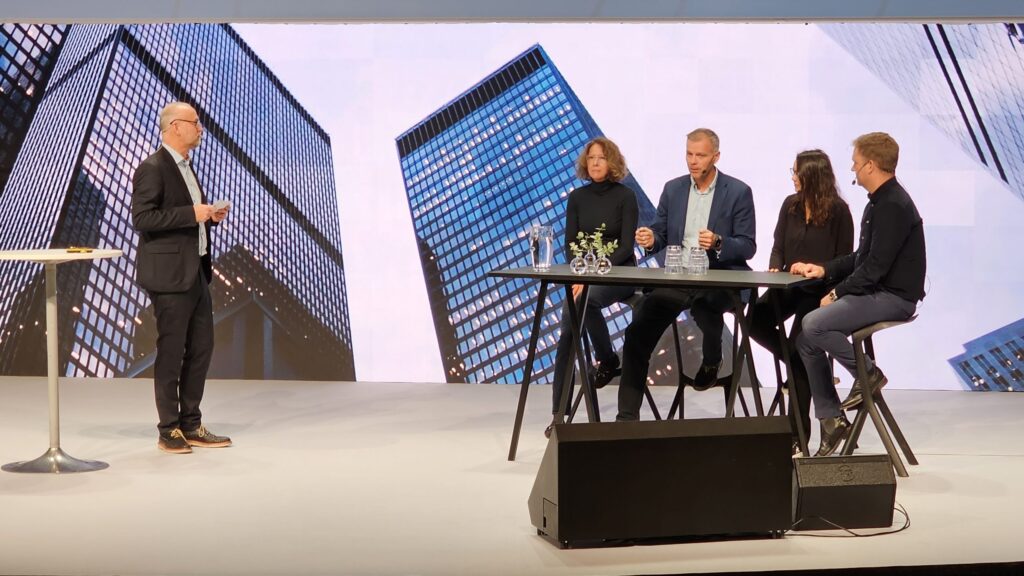 Four people sit at a tall table on stage, speaking, whilst one person stands beside them. A backdrop shows tall city buildings. The scene appears to be a panel discussion or conference.