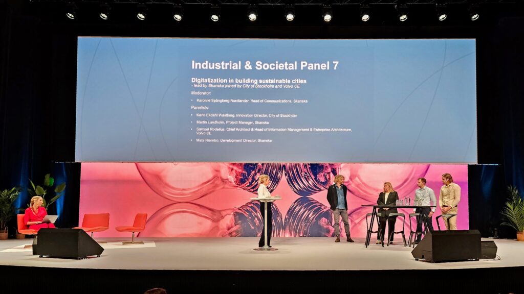 A panel of six people, five seated or standing at tables and one at a lectern, participate in a discussion on stage. A large screen above them displays Industrial & Societal Panel 7: Digitalisation in building sustainable cities.