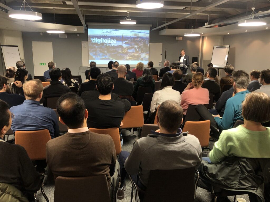 A group of people sit in rows facing a presenter at the front of a conference room, watching a presentation titled digitalise. Some attendees take notes; the room appears professional and modern.