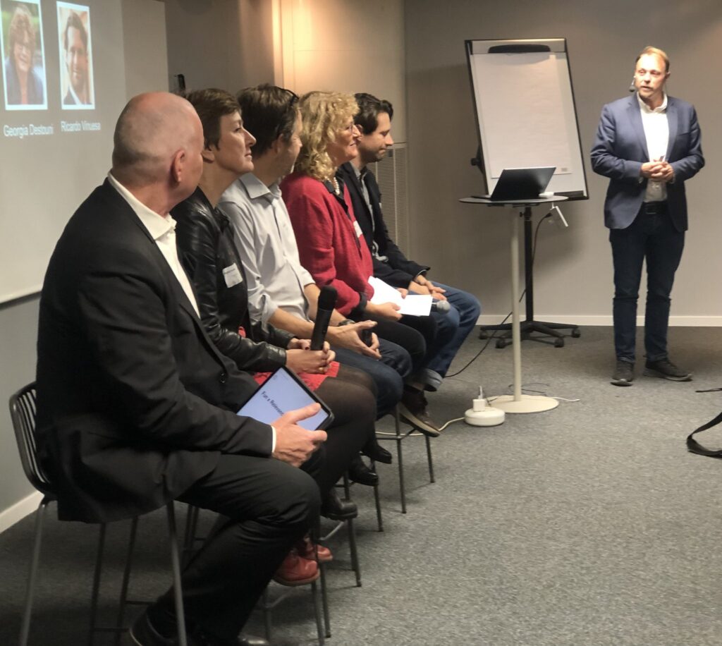 A group of six people sit in a row facing an audience, while a man stands near a flipchart and laptop, presenting in a conference room setting.