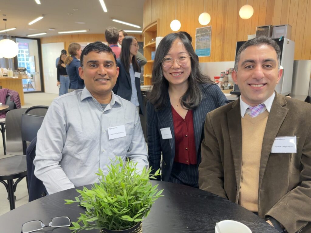 Three people wearing name badges smile while sitting at a round table in a modern, well-lit room with wooden walls. A small green plant is on the table in front of them, and others are in the background.