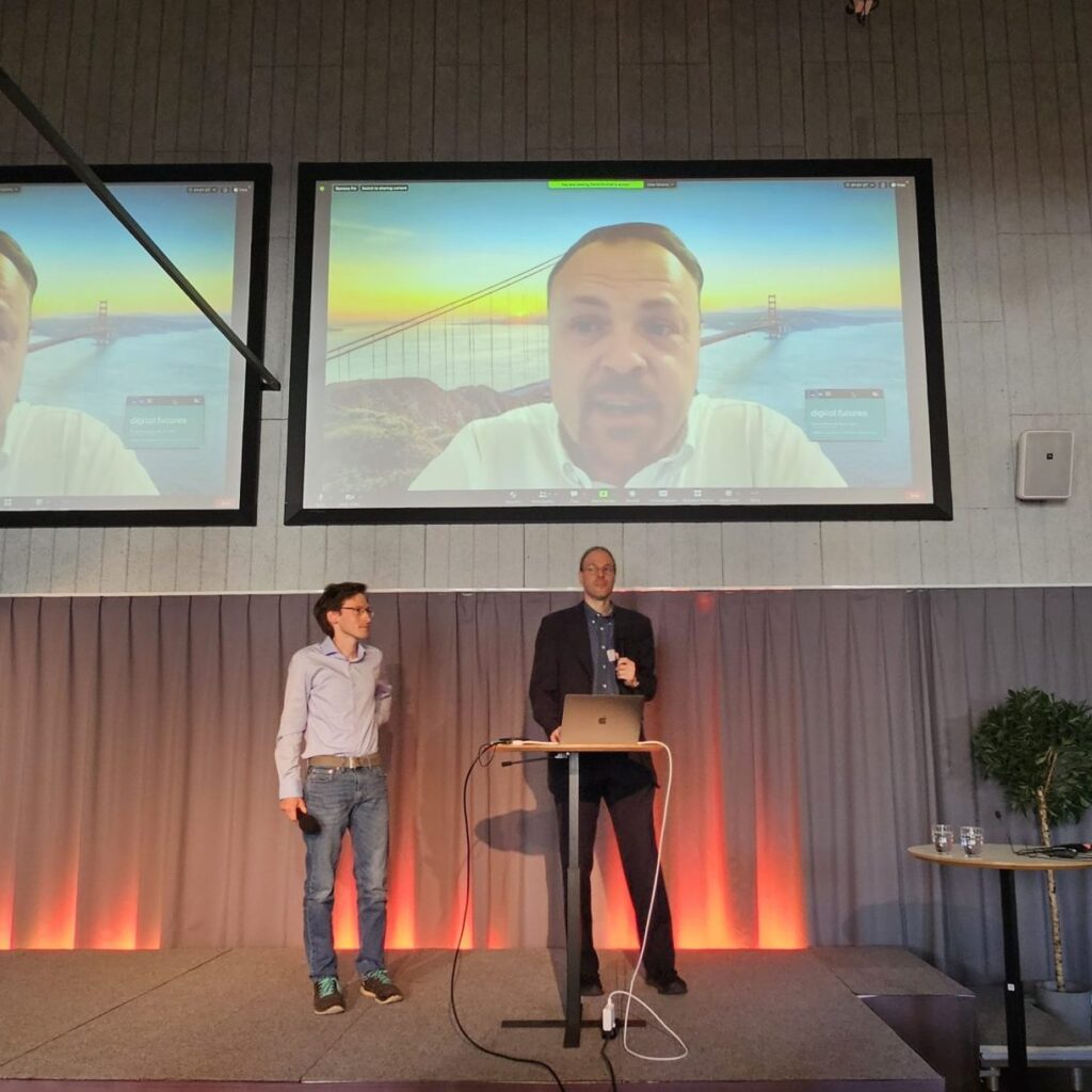 Two men stand on a stage with microphones and a laptop, whilst a large screen behind them shows a man on a video call with the Golden Gate Bridge as his virtual background.