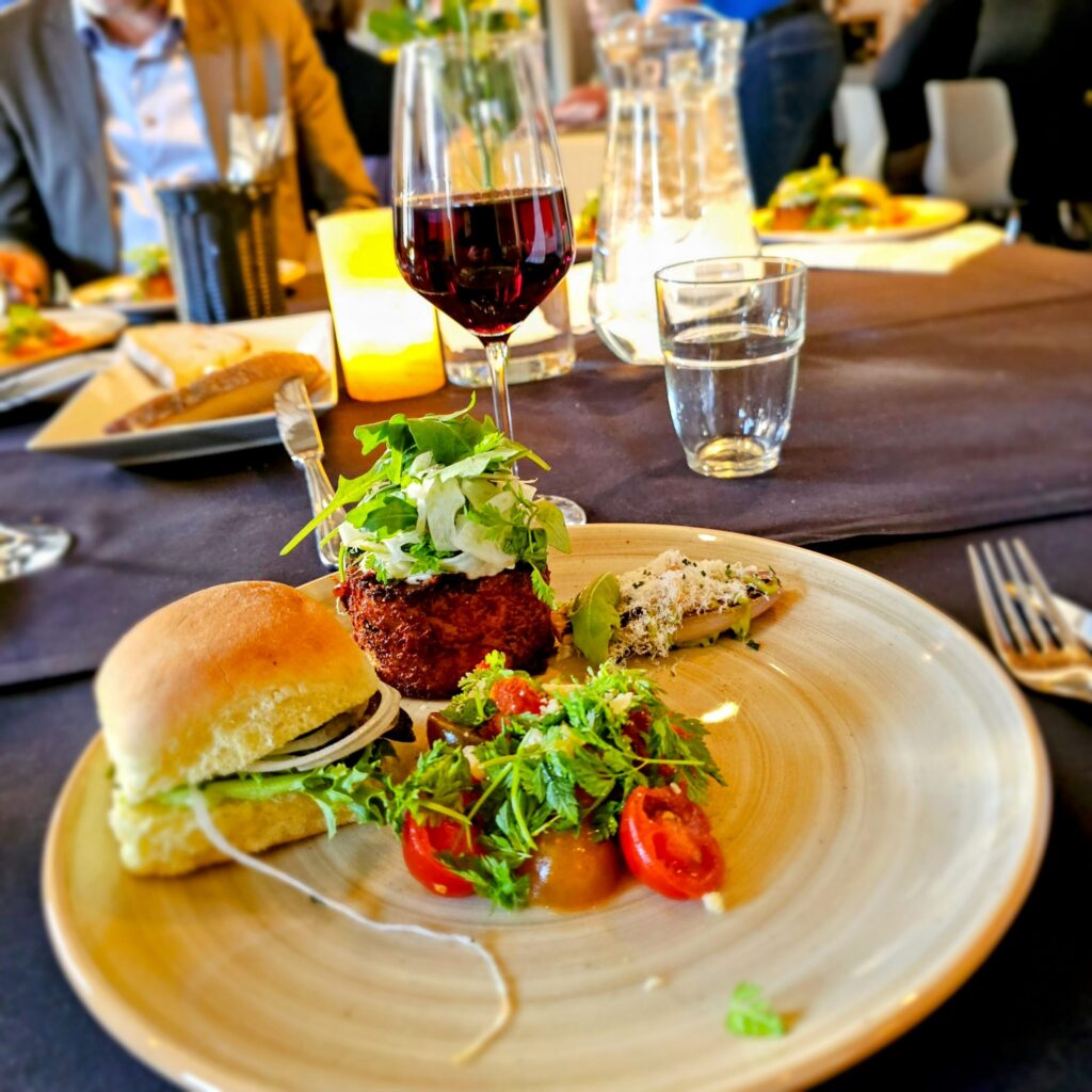 A plated meal features a slider, a salad with cherry tomatoes, and a garnished side, set on a table with a glass of red wine, water, and candles, with people dining in the background.