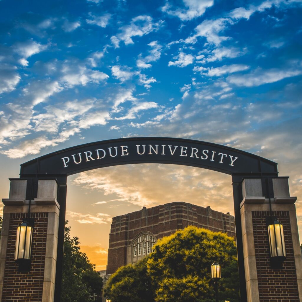 An archway with Purdue University written on it stands in front of a brick building, with trees and lanterns on either side, set against a vibrant sunset sky with clouds.