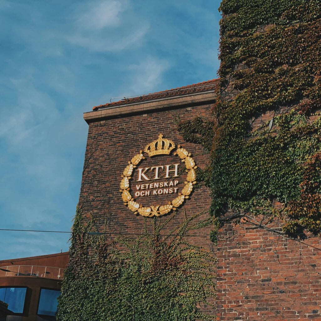 A brick building with creeping ivy displays a golden KTH Vetenskap och Konst emblem and crown under a blue sky, representing the Royal Institute of Technology in Stockholm.