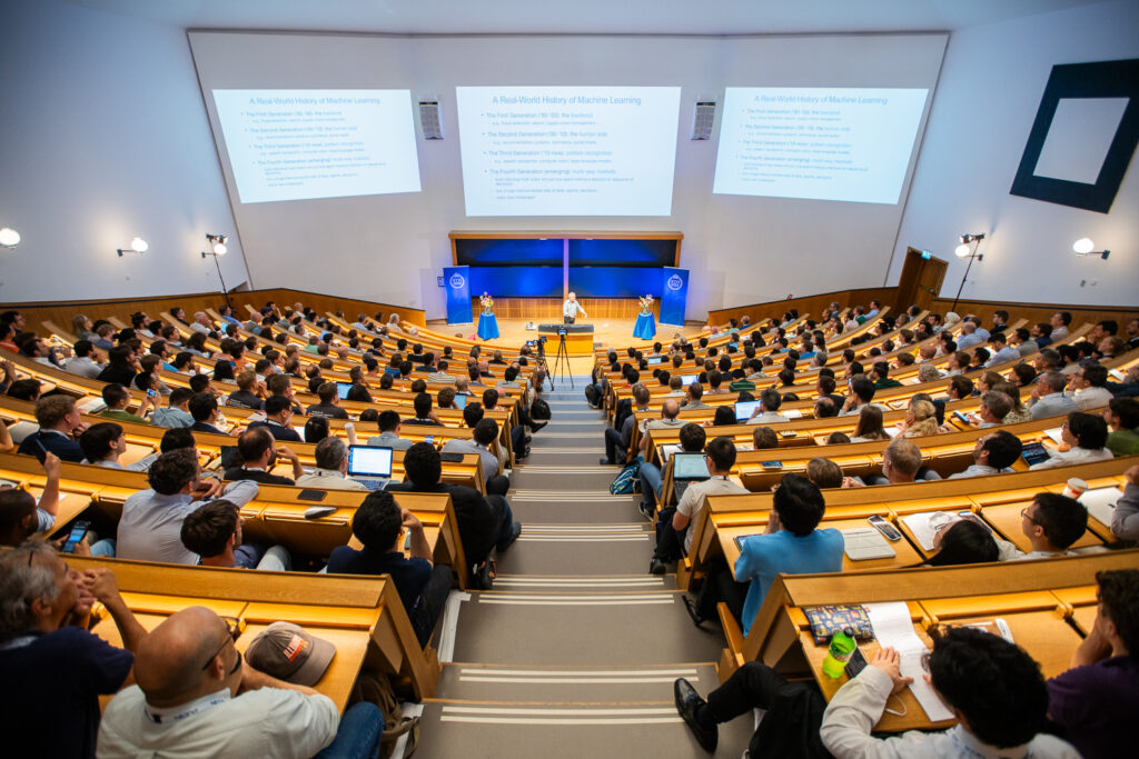 A large lecture theatre filled with people listening to three presenters on stage, with slides about machine learning projected on two screens at the front of the room.