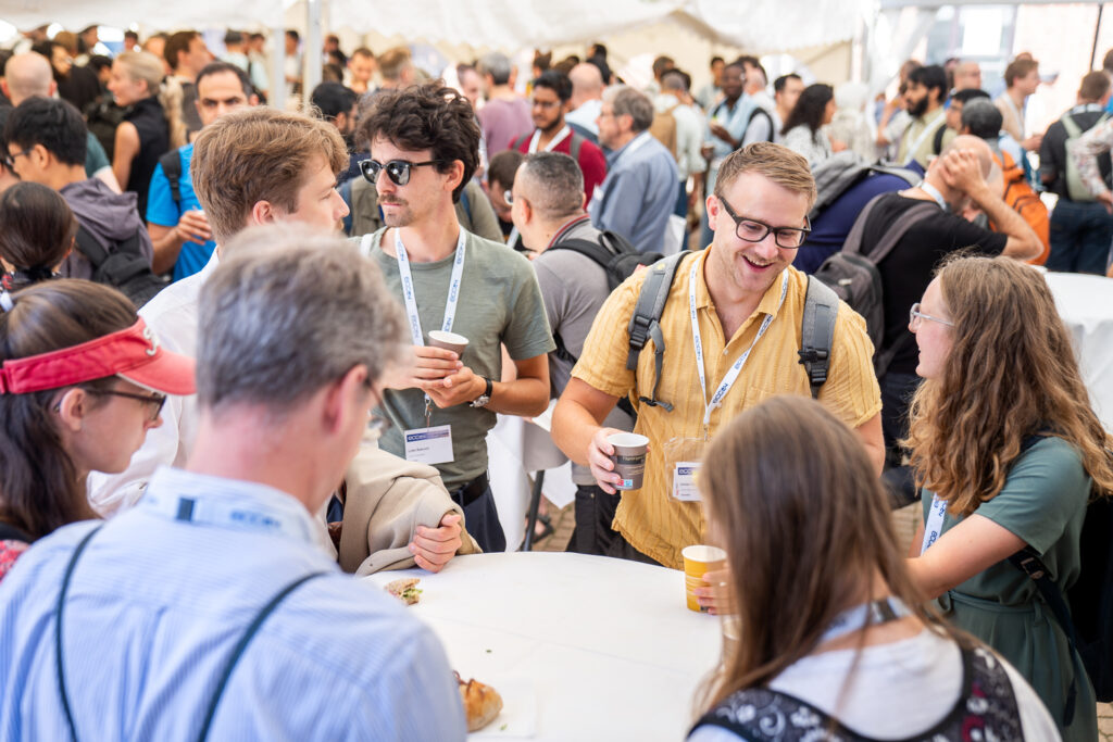 A large group of people, many with name badges, socialise around tables at a busy networking event. Some hold coffee cups and engage in lively conversation under a white marquee.
