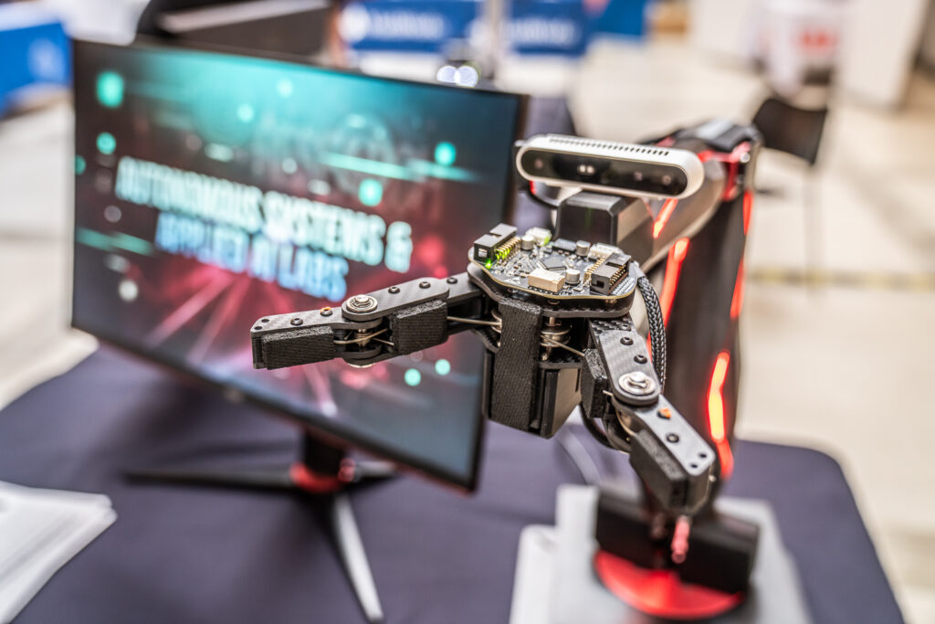 A robotic arm with a gripper is positioned near a computer monitor displaying the text AUTONOMOUS SYSTEMS & AI EXPLORATION LABS in a bright, futuristic style. The setting appears to be a tech lab or expo.
