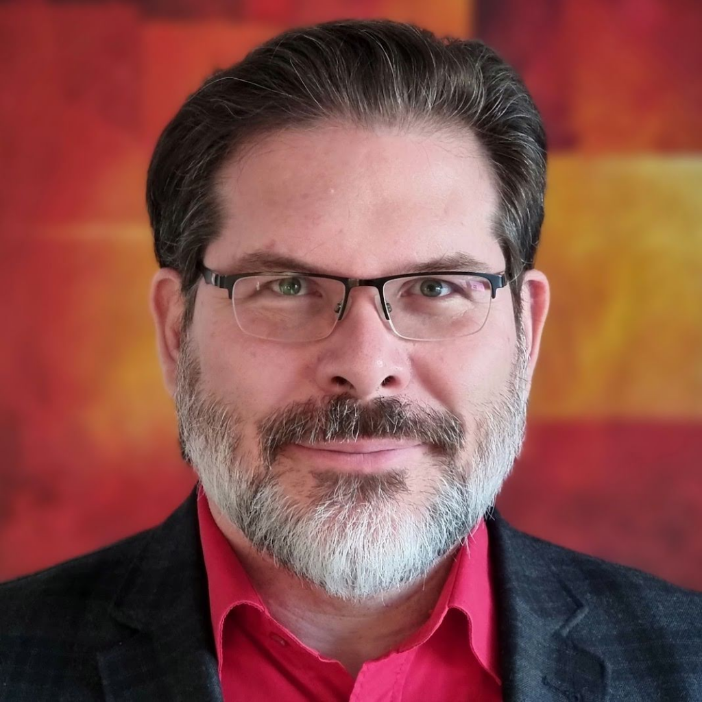 A man with short dark hair, a salt-and-pepper beard, and glasses, wearing a red shirt and dark blazer, stands in front of a blurred red and yellow background.